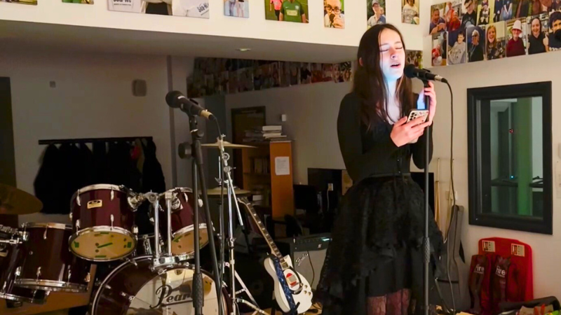 Young woman singing at mic, she is holding a mobile phone and there is a drumkit behind her