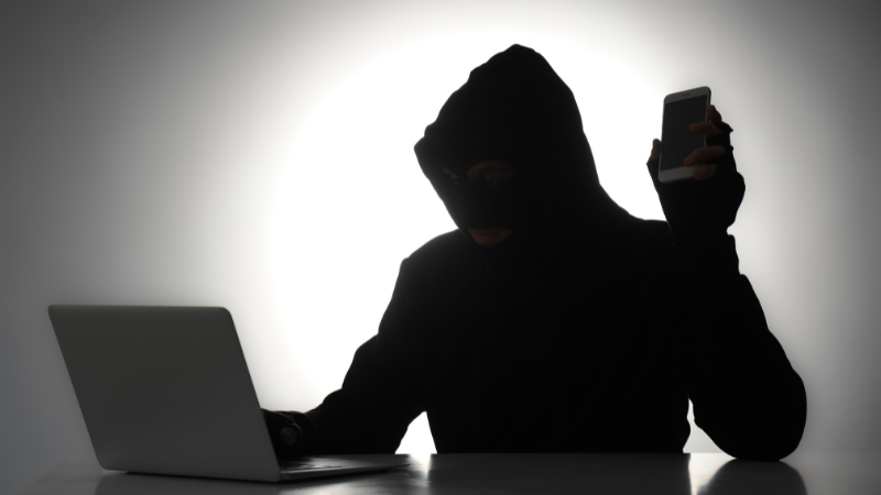 Person wearing a hoodie in silhouette on a laptop and holding up a mobile phone